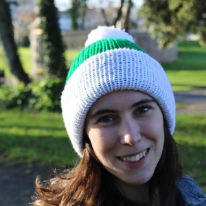 May include: A white knitted beanie with green stripes and a white pom pom on top. The beanie is being worn by a person with long brown hair.