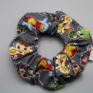Puede incluir: Scrunchie gris con un patrón de superhéroes de dibujos animados en varias poses. El scrunchie tiene una pequeña etiqueta blanca con el texto "Little Bits" en ella.