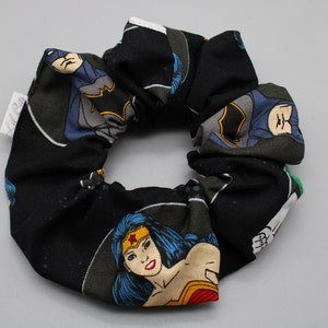 Puede incluir: Scrunchie negro con temática de superhéroes. El scrunchie presenta a Wonder Woman, Batman y Superman en un estilo de cómic.
