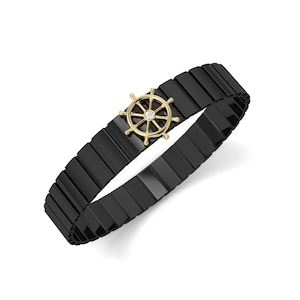 May include: Black bracelet with a gold-colored ship wheel charm. The bracelet is made of rectangular black links. The ship wheel charm has a small, clear stone in the center. The bracelet is shown on a white background.