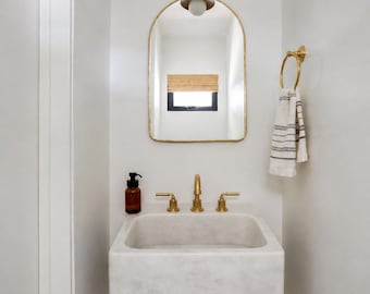 Op de afbeelding: Een moderne badkamer met een marmeren wastafel, gouden kranen en een bijpassende boogvormige spiegel. Een gestreepte handdoek hangt aan een gouden handdoekring. Wandverlichting en een zeepdispenser maken het minimalistische ontwerp compleet.