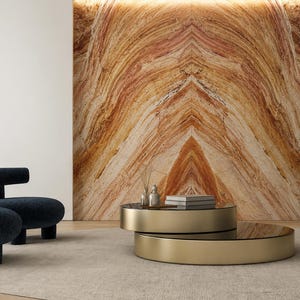 Op de afbeelding: Een moderne woonkamer met een gelaagde gouden salontafel met een glazen blad, een blauwe stoel met textuur en een grote, bewerkte stenen muur. De kamer heeft een neutraal tapijt en een boekenkast.