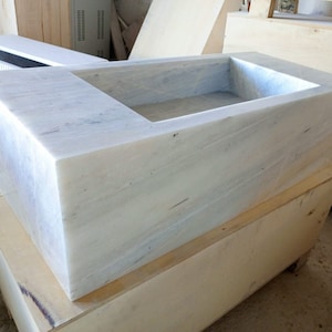 May include: A white marble sink with a rectangular shape and a square basin. The sink is sitting on a wooden crate.