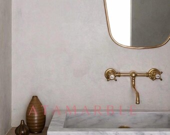 May include: A rectangular white marble sink with gray veining sits on a light wood counter. A wall-mounted brass faucet with two cross handles is positioned above the sink. An oval mirror with a thin brass frame hangs above the faucet.