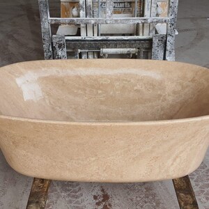 May include: A large, oval-shaped bathtub made of light brown travertine stone. The bathtub is sitting on a concrete floor.