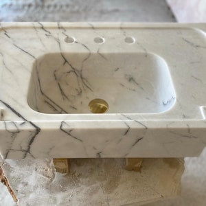 May include: White marble bathroom sink with a rectangular shape and three holes for faucets. The sink is sitting on wooden blocks.