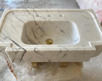 May include: White marble bathroom sink with a rectangular shape and three holes for faucets. The sink is sitting on wooden blocks.
