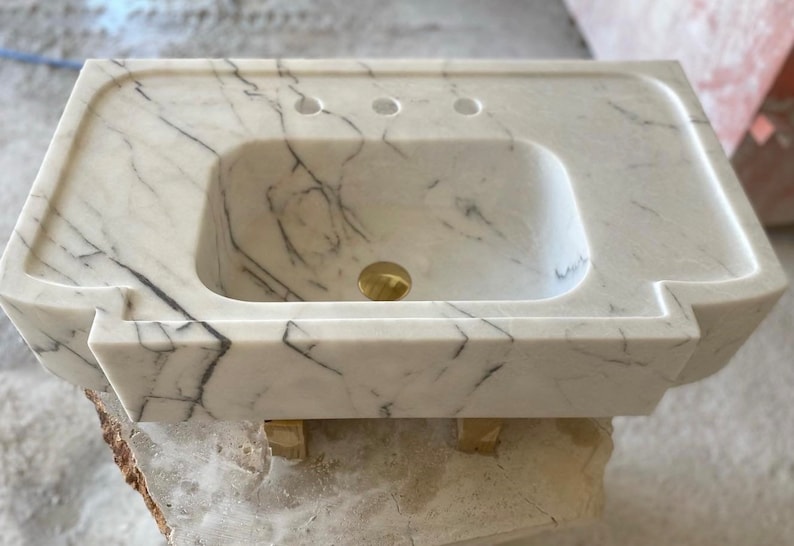 Puede incluir: Lavabo de ba&ntilde;o de m&aacute;rmol blanco con forma rectangular y tres agujeros para grifos. El lavabo est&aacute; sobre bloques de madera.