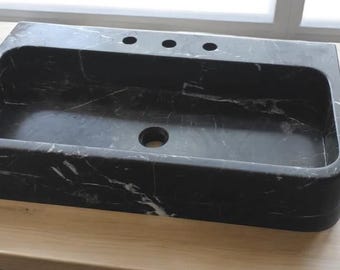 May include: A black marble vessel sink with a rectangular shape and a rounded front edge. The sink has three holes for faucets and a drain hole in the center.
