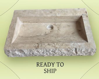 May include: A rectangular, beige stone sink with a central drain hole. The sink has a rough, textured exterior and a smooth interior basin. The words "READY TO SHIP" are printed below the sink.