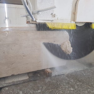 May include: A large block of beige stone being cut by a circular saw with water being sprayed on the blade. The saw is attached to a white machine with yellow brushes.