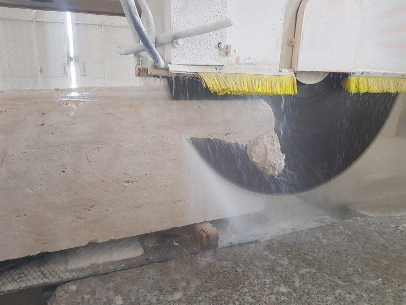 May include: A large block of beige stone being cut by a circular saw with water being sprayed on the blade. The saw is attached to a white machine with yellow brushes.
