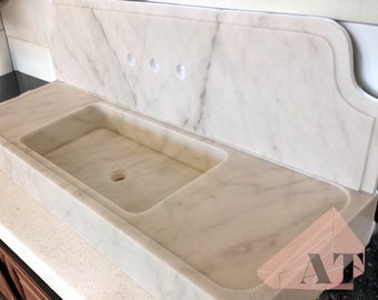 Può includere: Un lavandino in marmo bianco con un foro di scarico singolo e uno schienale con tre fori. Il lavandino è incassato in un piano di lavoro e ha un bordo curvo.