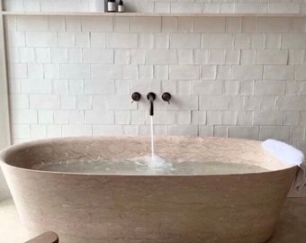 May include: A large, oval-shaped bathtub made of natural stone with a white towel on the edge. The bathtub is filled with water and a faucet is running, filling the tub.