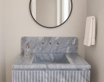 May include: A gray marble bathroom vanity with a rectangular sink and vertical grooved detailing. A round black mirror and a two-light fixture are above the vanity. A white towel hangs on the wall.