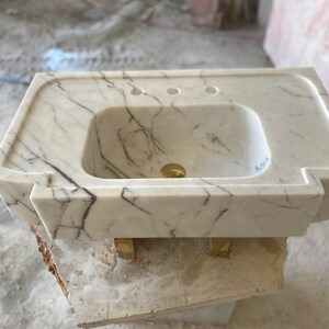 Puede incluir: Lavabo de ba&ntilde;o de m&aacute;rmol blanco con forma rectangular y un borde frontal curvo. El lavabo tiene tres agujeros para grifos y un desag&uuml;e.
