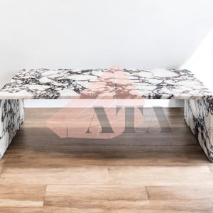 May include: A rectangular marble coffee table with a white base and black veining. The table has a solid, block-like design with a polished surface. The table is set on a wooden floor.