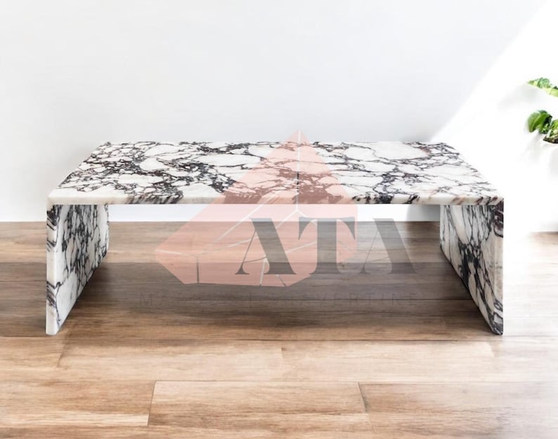 May include: A rectangular marble coffee table with a white base and black veining. The table has a solid, block-like design with a polished surface. The table is set on a wooden floor.