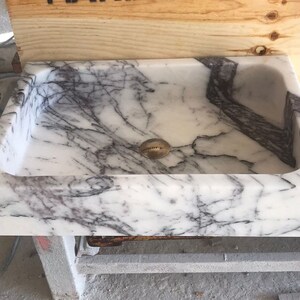 May include: A white marble sink with gray veining. The sink has a rounded rectangular shape and a drain hole in the center.