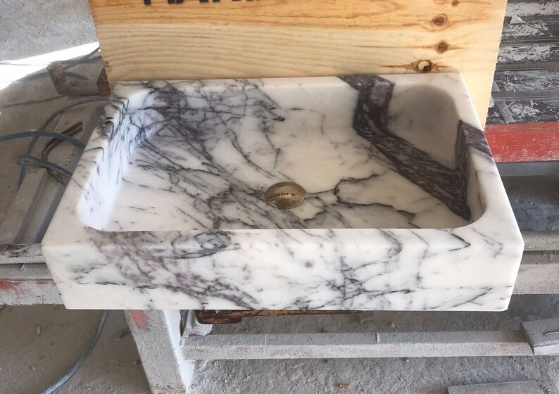 May include: A white marble sink with gray veining. The sink has a rounded rectangular shape and a drain hole in the center.