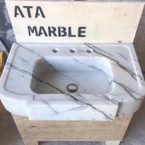 Puede incluir: Un lavabo de ba&ntilde;o de m&aacute;rmol blanco con tres agujeros para grifos. El lavabo est&aacute; montado sobre una base de madera y tiene un borde delantero curvo. El lavabo est&aacute; etiquetado como "ATA MARBLE".