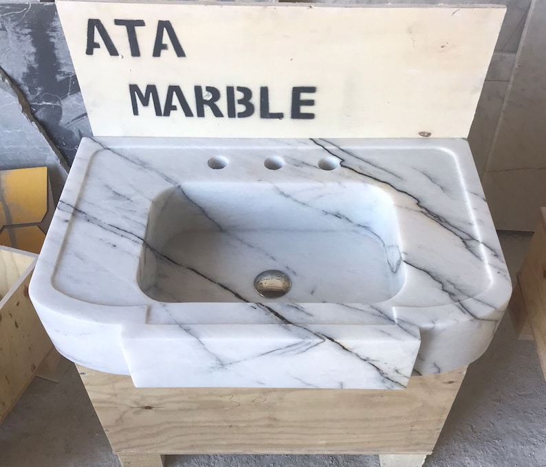 Puede incluir: Un lavabo de ba&ntilde;o de m&aacute;rmol blanco con tres agujeros para grifos. El lavabo est&aacute; montado sobre una base de madera y tiene un borde delantero curvo. El lavabo est&aacute; etiquetado como "ATA MARBLE".