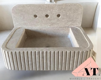 May include: A rectangular, light gray marble sink with a backsplash featuring three faucet holes. The sink basin is rectangular with a drain. The front of the sink has vertical, grooved detailing. The logo "ATA MARBLE | TRAVERTINE" is in the bottom right corner.