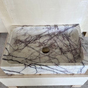 May include: A white marble sink with purple veins. The sink is rectangular and has a round drain hole in the center.