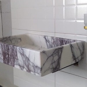 May include: A white marble sink with purple veins, mounted on a white tiled wall. The sink is rectangular and has a slightly rounded edge.