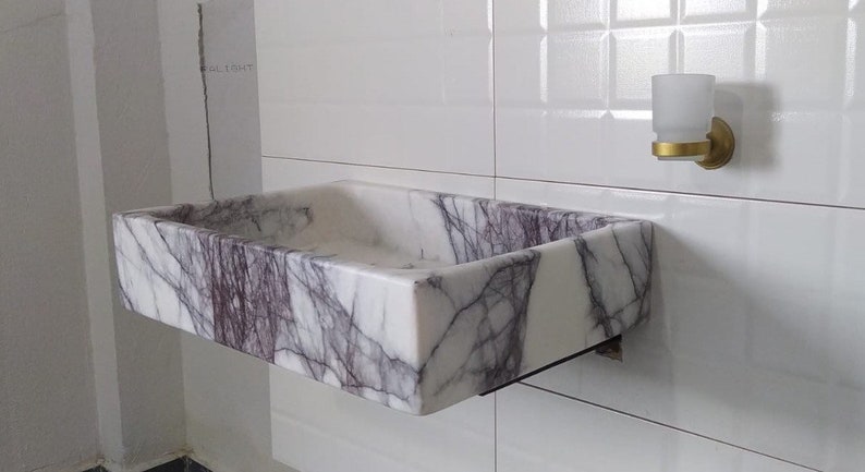 May include: A white marble sink with purple veins, mounted on a white tiled wall. The sink is rectangular and has a slightly rounded edge.