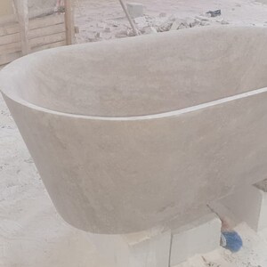 May include: A large, oval-shaped bathtub made of light gray stone. The bathtub is sitting on blocks of wood and is being worked on by a craftsman.