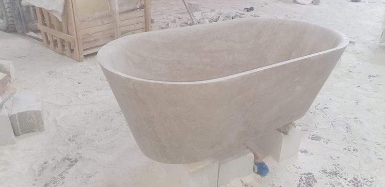 May include: A large, oval-shaped bathtub made of light gray stone. The bathtub is sitting on blocks of wood and is being worked on by a craftsman.