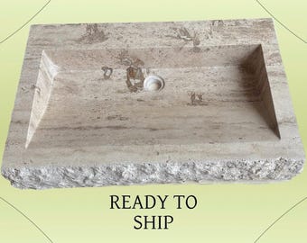 May include: A rectangular, beige stone sink with a recessed basin and a central drain hole. The sink has a rough, textured exterior and a smooth interior. The words "READY TO SHIP" are printed below the sink.