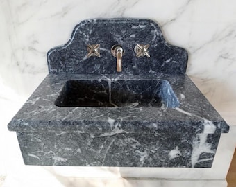 Puede incluir: Un lavabo de mármol gris oscuro con una cuenca rectangular y una parte superior arqueada decorativa. El lavabo tiene dos grifos cromados con manijas en forma de cruz.