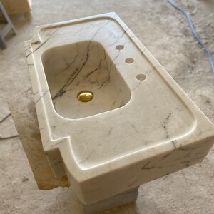 Puede incluir: Un lavabo de ba&ntilde;o de m&aacute;rmol blanco con un solo agujero para un grifo y tres agujeros para accesorios. El lavabo est&aacute; montado sobre una base de hormig&oacute;n.