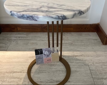 May include: A round white marble table top with grey veining sits on a gold metal base with a circular design. The table is on a light wood floor.
