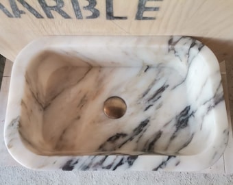 May include: White marble sink with a gray veining pattern. The sink has a round drain hole and a silver drain stopper.