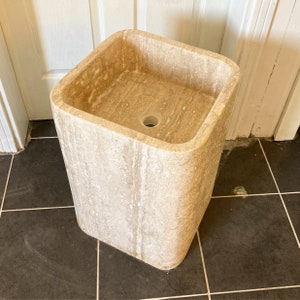 May include: A square, freestanding stone sink with a light beige color and a smooth, rounded edge. The sink is made of travertine and has a drain hole in the center.