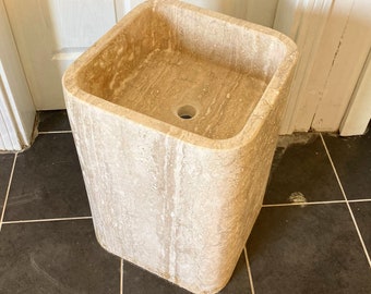 May include: A square, freestanding stone sink with a light beige color and a smooth, rounded edge. The sink is made of travertine and has a drain hole in the center.