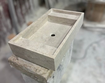Puede incluir: Un lavabo rectangular de piedra con un solo agujero de drenaje y un pequeño estante en un lado. El lavabo está hecho de una piedra gris clara con una textura rugosa.