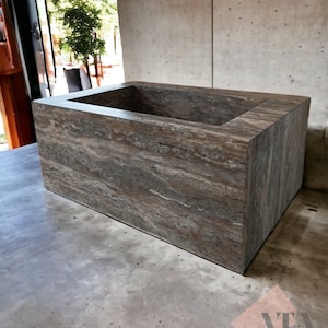 May include: A rectangular, brown stone bathtub with a textured surface. The tub has a recessed interior and is set on a concrete surface. The image is taken in natural light, highlighting the stone's natural grain and color variations. The product is a bathroom fixture.