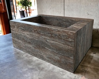 May include: A rectangular, brown stone bathtub with a textured surface. The tub has a recessed interior and is set on a concrete surface. The image is taken in natural light, highlighting the stone's natural grain and color variations. The product is a bathroom fixture.