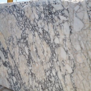 May include: Large slabs of marble with a complex pattern of white, gray, and black veining. The stone has a polished surface and a natural, organic appearance. The marble is stacked, showing the variation in the stone's design.