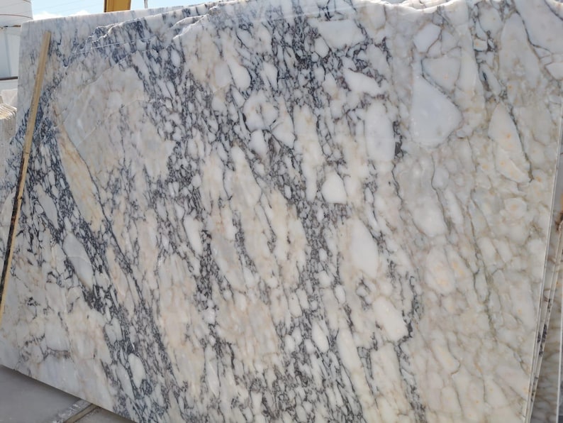 May include: Large slabs of marble with a complex pattern of white, gray, and black veining. The stone has a polished surface and a natural, organic appearance. The marble is stacked, showing the variation in the stone's design.