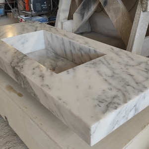 Puede incluir: Una encimera de m&aacute;rmol blanco con un corte rectangular para un lavabo. La encimera est&aacute; apoyada sobre una base de madera.