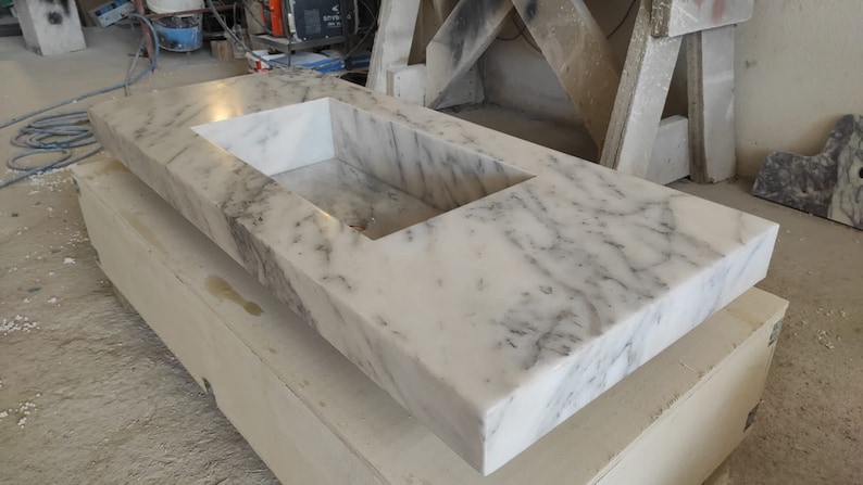 Puede incluir: Una encimera de m&aacute;rmol blanco con un corte rectangular para un lavabo. La encimera est&aacute; apoyada sobre una base de madera.