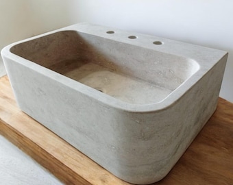 May include: A rectangular, off-white stone vessel sink with three drain holes. The sink is sitting on a wooden countertop.