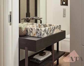 Puede incluir: Un lavabo de baño de mármol blanco con vetas negras se asienta sobre un tocador de madera oscura con un marco de metal. El lavabo tiene un grifo de níquel cepillado y una pequeña planta en una cesta tejida.