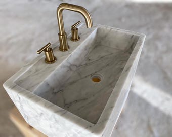 May include: A white marble bathroom sink with a rectangular shape and a single drain hole. The sink has three gold faucets.