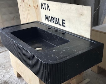 May include: A black marble bathroom sink with a rectangular basin and a decorative fluted edge. The sink is mounted on a wooden base and has three holes for faucets. The sink is labeled "ATA MARBLE".